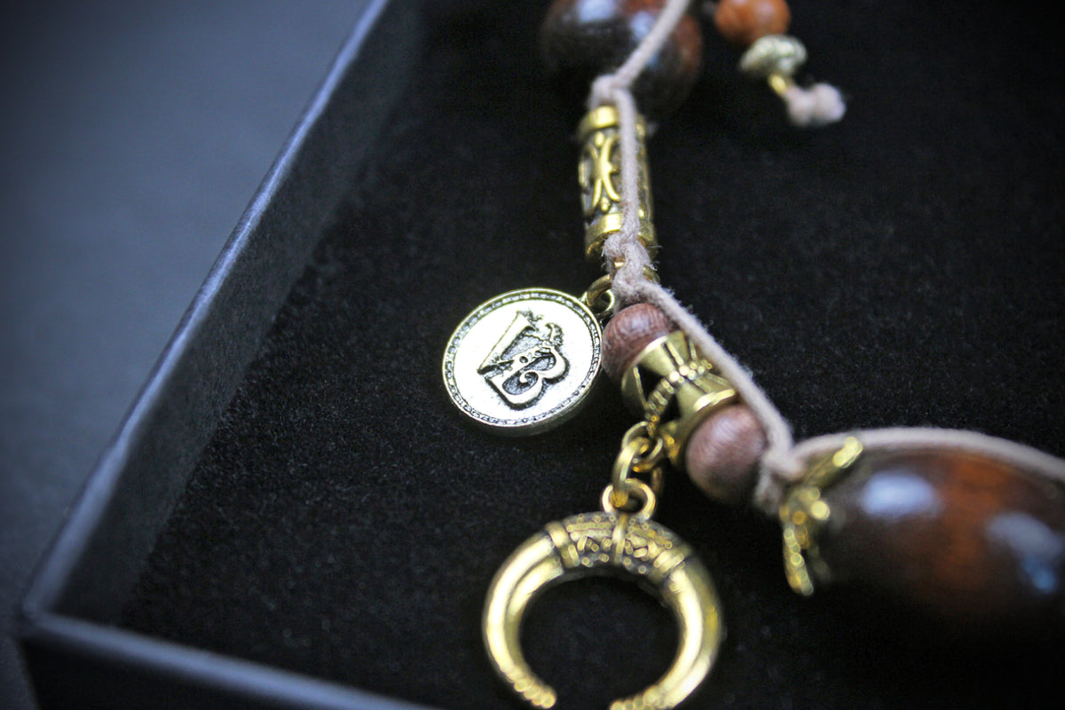 A close-up of a beaded necklace or bracelet with metallic gold-toned charms, including a circular pendant with an engraved symbol and a crescent moon charm, resting in a black presentation box.