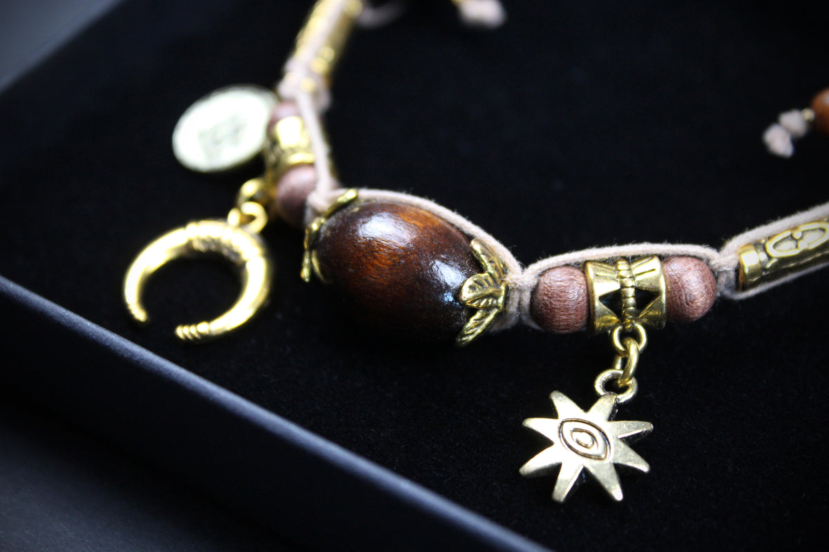 A bohemian-style beaded bracelet with a large polished dark brown wooden bead, pinkish-brown beads, gold-toned metal charms including a crescent moon, a star, and a cylindrical accent, displayed on a black velvet jewelry box.
