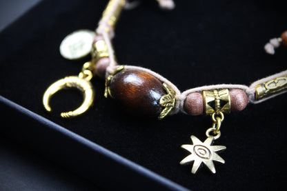 A bohemian-style beaded bracelet with a large polished dark brown wooden bead, pinkish-brown beads, gold-toned metal charms including a crescent moon, a star, and a cylindrical accent, displayed on a black velvet jewelry box.