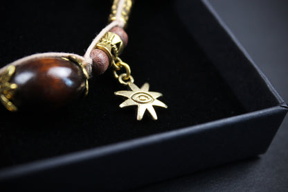A close-up of a beaded necklace with a polished dark brown bead, gold-toned metal caps, and a small gold-toned starburst charm, displayed inside a black presentation box.