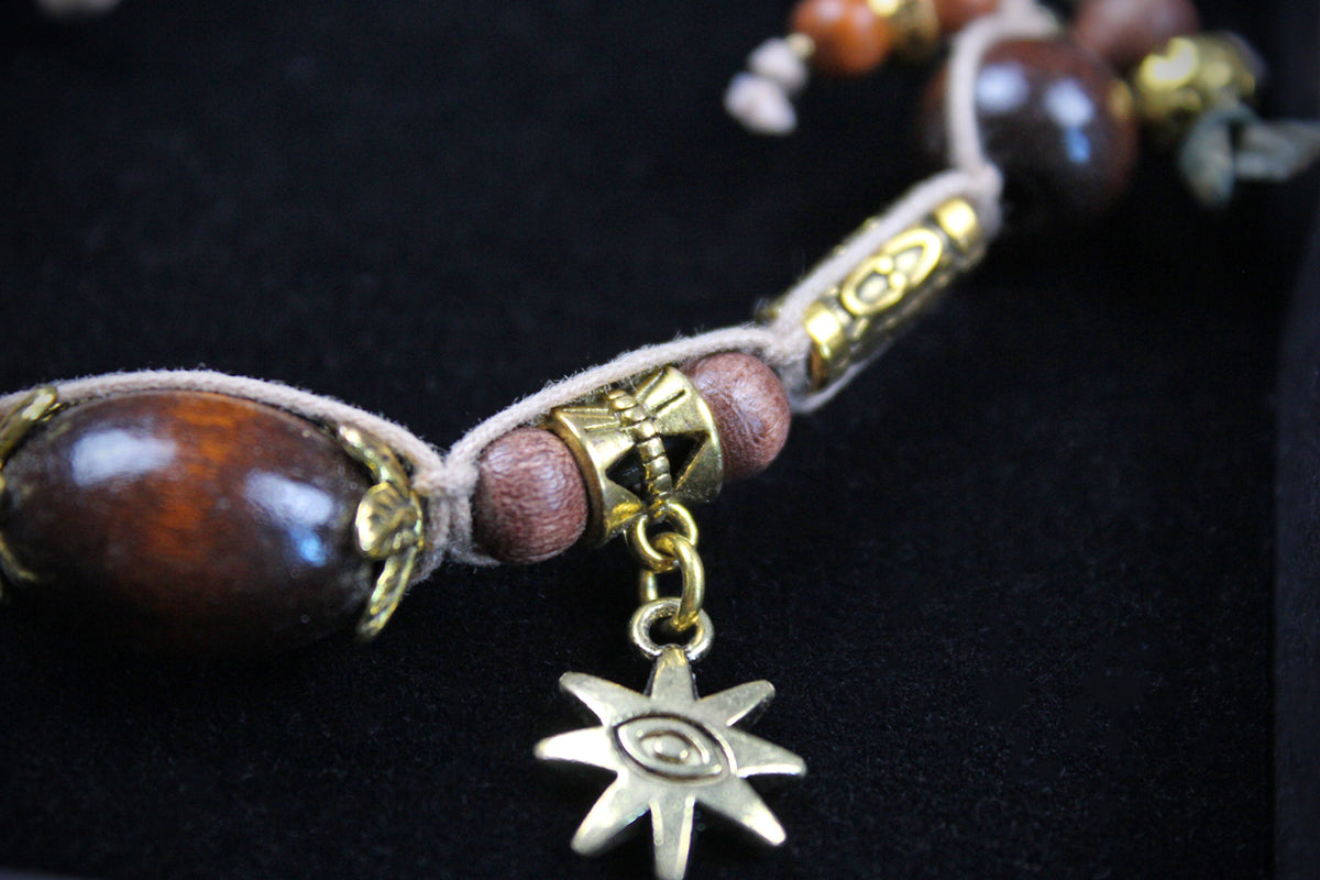 A close-up of a beaded cord accessory with large polished dark brown wooden beads, smaller reddish-brown beads, gold-toned metal spacers, and a dangling gold-toned star charm with an eye motif, set against a black background.