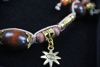 A close-up of a beaded cord accessory with large polished dark brown wooden beads, smaller reddish-brown beads, gold-toned metal spacers, and a dangling gold-toned star charm with an eye motif, set against a black background.