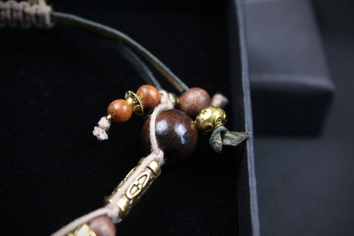 A close-up of a beaded cord accessory with dark brown wooden beads, small metallic gold-toned beads, and a dark green cord, resting on a black surface.