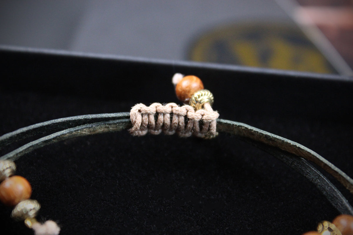 A close-up of a beaded bracelet with a dark green cord, a beige woven knot detail, and brown wooden beads with gold-toned spacer accents, resting on a black velvet surface.