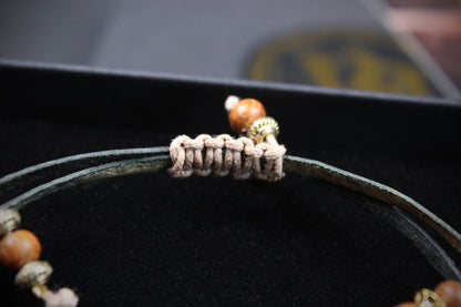 A close-up of a beaded bracelet with a dark green cord, a beige woven knot detail, and brown wooden beads with gold-toned spacer accents, resting on a black velvet surface.