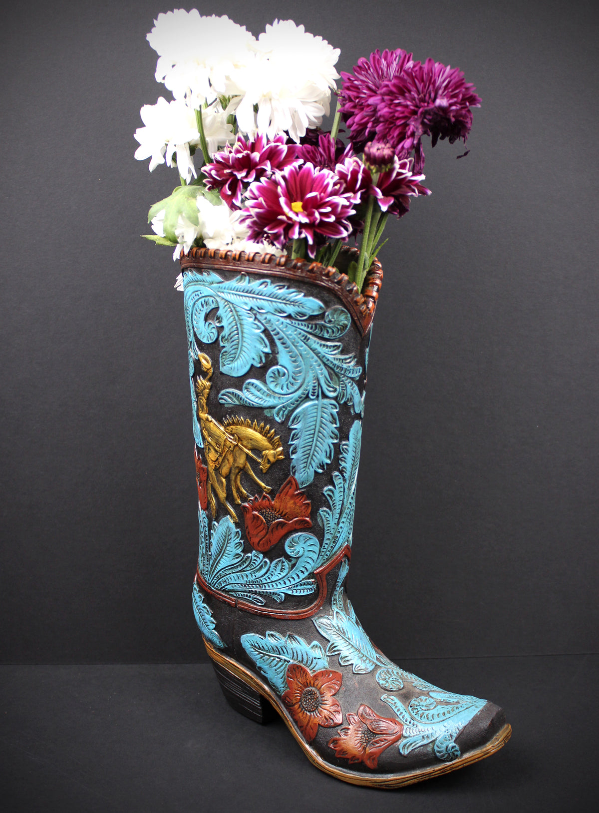 A decorative cowboy boot vase filled with white and purple flowers, set against a dark gray background. The boot features intricate turquoise, brown, and gold floral and western embroidery.