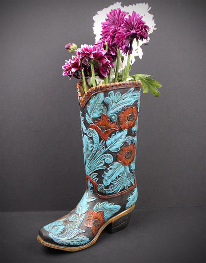 A decorative cowboy boot vase filled with purple flowers, featuring turquoise and rust floral tooled leather on a dark brown background, set against a dark gray backdrop.