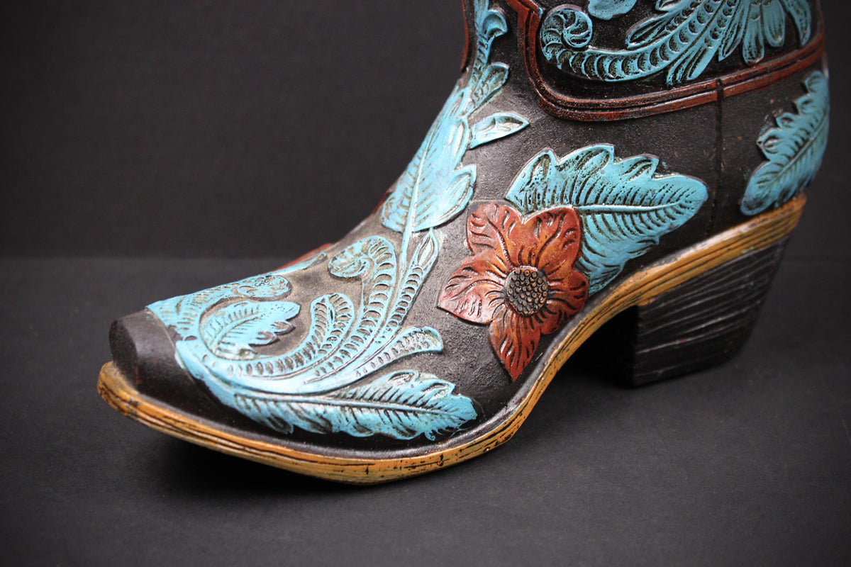 A close-up of a hand-tooled leather cowboy boot with turquoise blue floral and leaf designs, a reddish-brown flower, and a dark brown heel on a dark gray background.