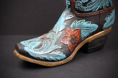 A close-up of a hand-tooled leather cowboy boot with turquoise blue floral and leaf designs, a reddish-brown flower, and a dark brown heel on a dark gray background.