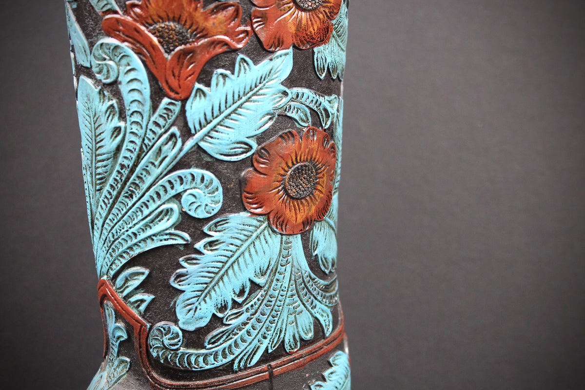 Close-up of a hand-tooled western boot shaft featuring embossed floral and leaf patterns in turquoise, reddish-brown, and dark brown against a dark background.