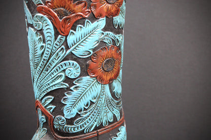 Close-up of a hand-tooled western boot shaft featuring embossed floral and leaf patterns in turquoise, reddish-brown, and dark brown against a dark background.