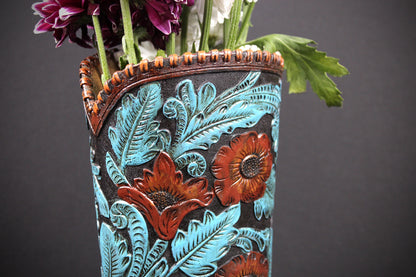 A hand-tooled leather flower vase with turquoise and red floral designs, filled with flowers, set against a dark gray background.