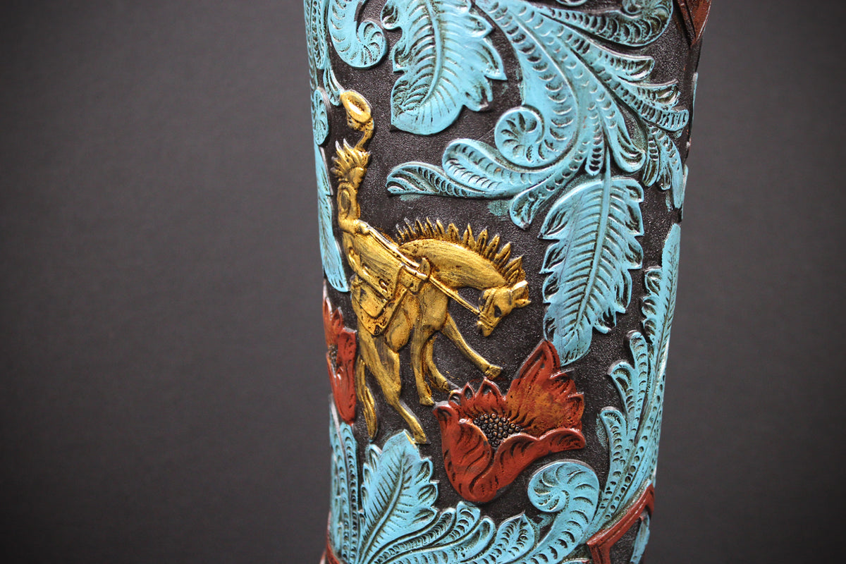 Close-up of a decorative cowboy boot shaft featuring turquoise blue embossed leaf patterns, a gold-toned cowboy riding a horse, and red floral accents on a dark brown background.