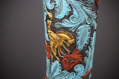 Close-up of a decorative cowboy boot shaft featuring turquoise blue embossed leaf patterns, a gold-toned cowboy riding a horse, and red floral accents on a dark brown background.