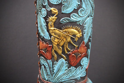 Close-up of a western-style boot featuring a black leather base with turquoise blue embossed floral and leaf patterns, a central gold-toned cowboy riding a horse, and red embossed flowers, set against a dark gray background.