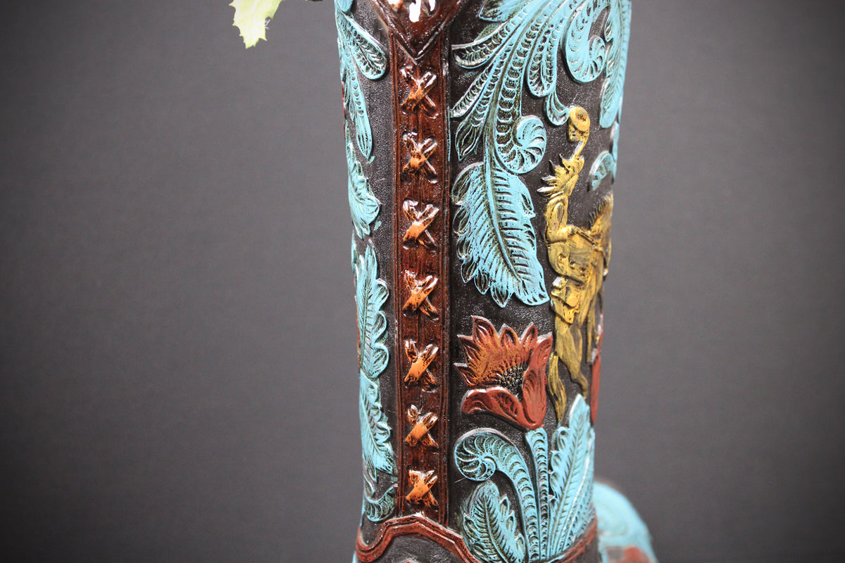 Close-up of a western boot shaft featuring turquoise blue foil, brown tooled leather, and gold and red floral embroidery against a dark background.
