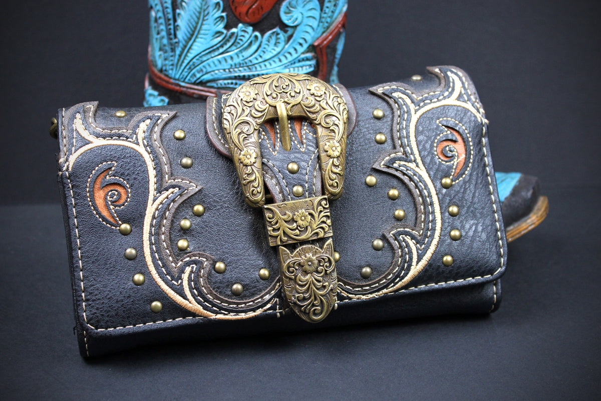 Close-up of a brown leather item with detailed embroidery and a bronze buckle centrepiece. The design features cut-out shapes, bronze studs, and a stitched border.