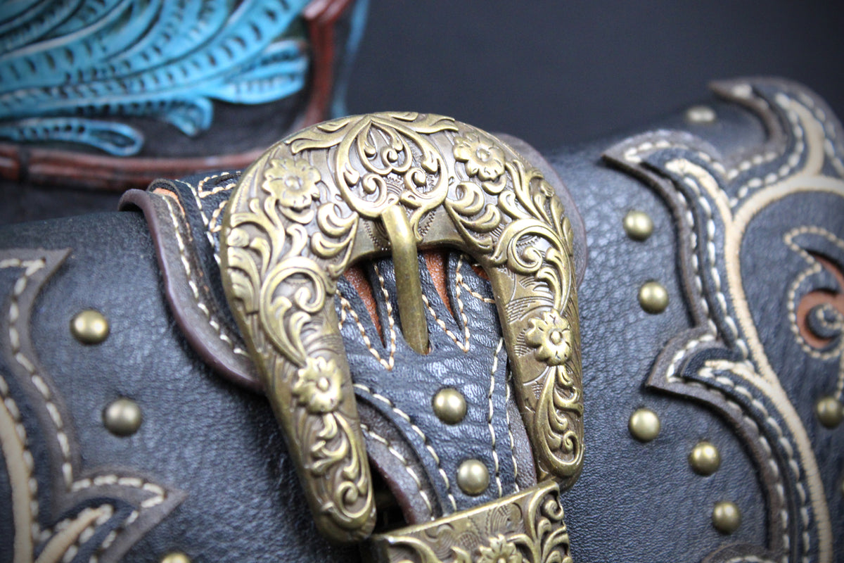 Close-up of a western leather saddle or horse blanket featuring an ornate brass buckle, dark brown leather panels, white contrast stitching, and brass studs.