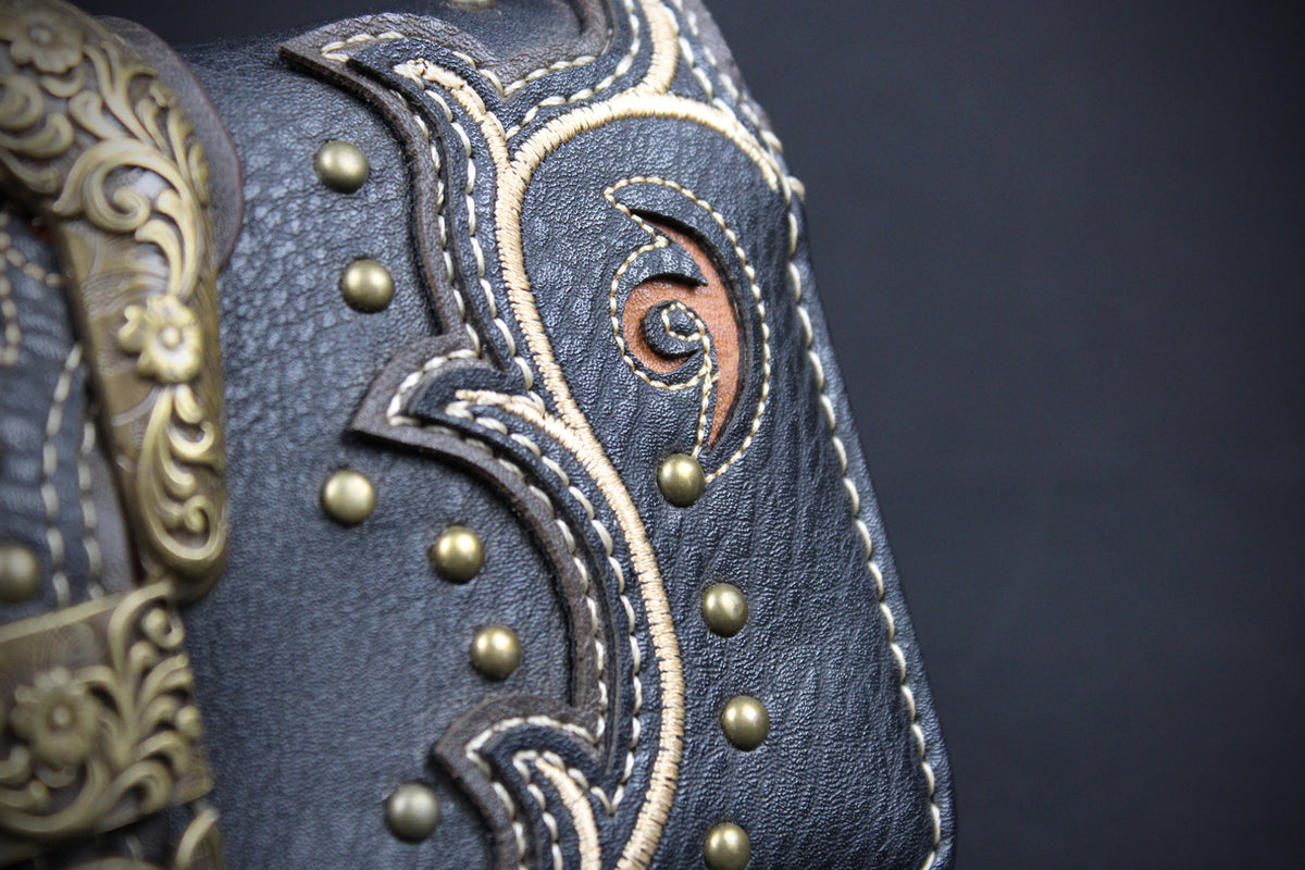 Close-up of a dark blue leather boot or mule featuring intricate western-style stitching, brass studs, and a decorative bronze-toned metal buckle on a dark background.
