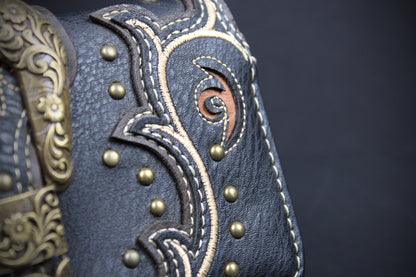 Close-up of a dark blue leather boot or mule featuring intricate western-style stitching, brass studs, and a decorative bronze-toned metal buckle on a dark background.