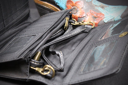 Close-up of an open black leather wallet or organizer with multiple card slots, a wrist strap, and gold-tone hardware, photographed against a dark background with a colorful boot partially visible.