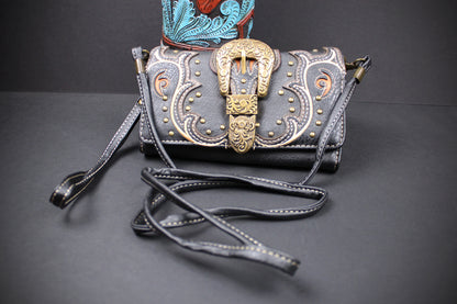 A black leather crossbody bag with an ornate western-style buckle closure, featuring gold-toned metal detailing and white contrast stitching, displayed on a dark gray surface.