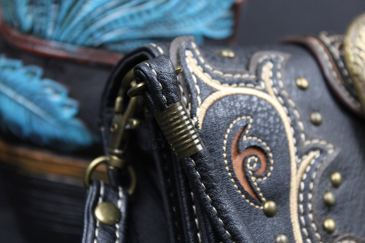 Close-up of a western-style black leather bag with gold-toned hardware, tan contrast stitching, and a carved paisley motif.
