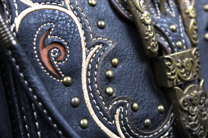 Close-up of a western-style leather bag or belt featuring dark navy leather, white contrast stitching, brass studs, and ornate antique bronze metal hardware.