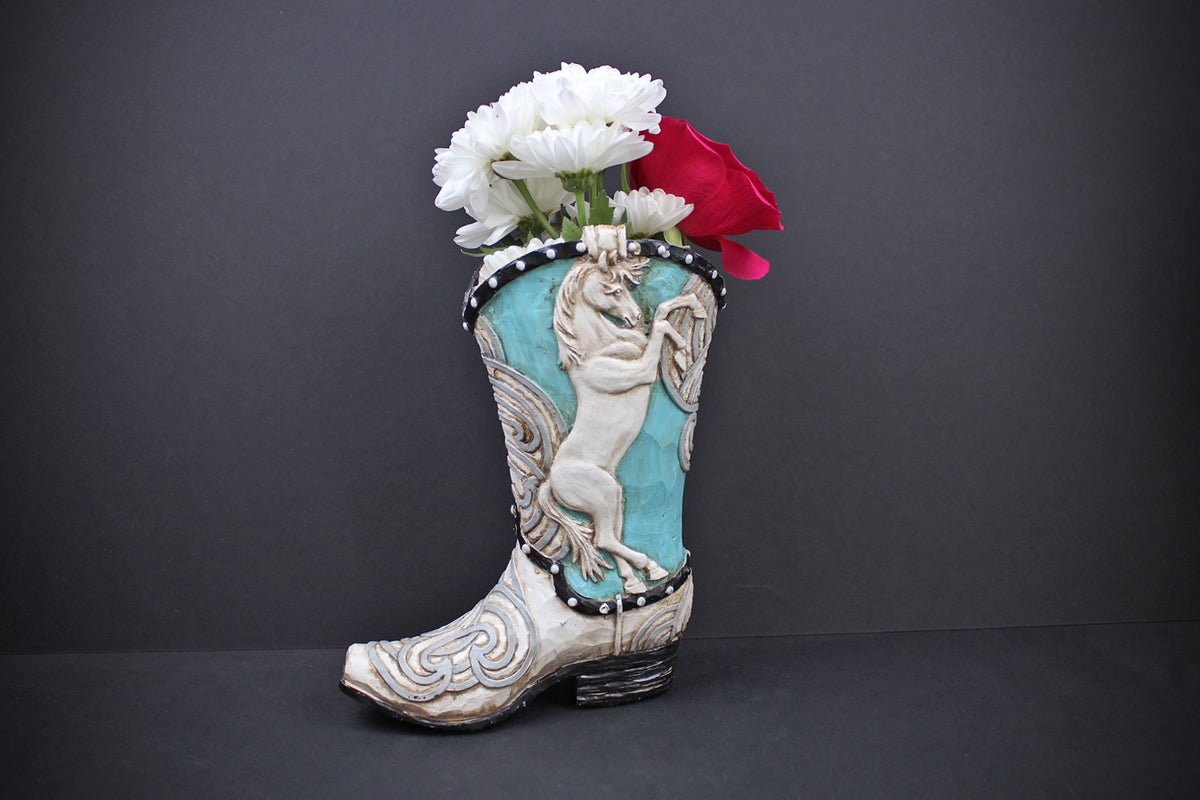 A ceramic cowboy boot planter with turquoise blue and white western detailing, filled with white flowers and a red rose, set against a dark gray background.