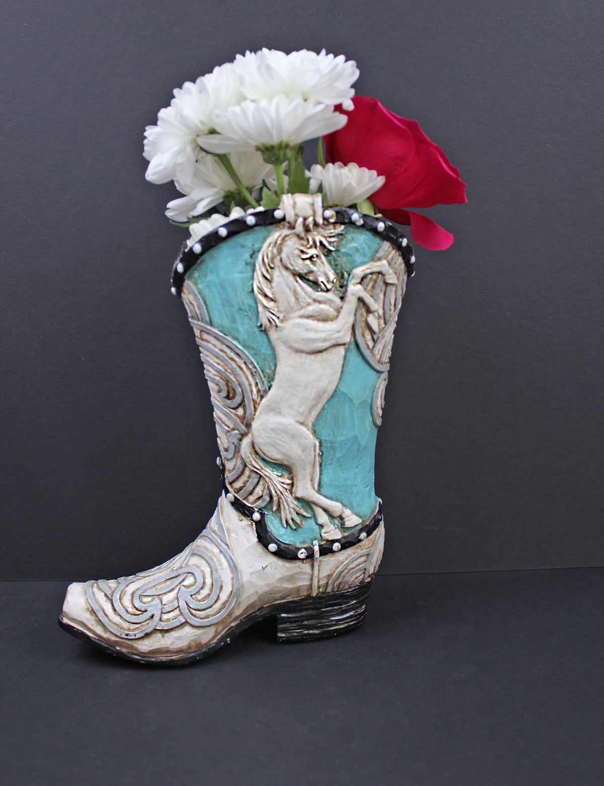 A ceramic cowboy boot flower vase with turquoise blue panels, a white prancing horse design, and white faux fur trim, filled with white flowers and a red rose against a dark gray background.