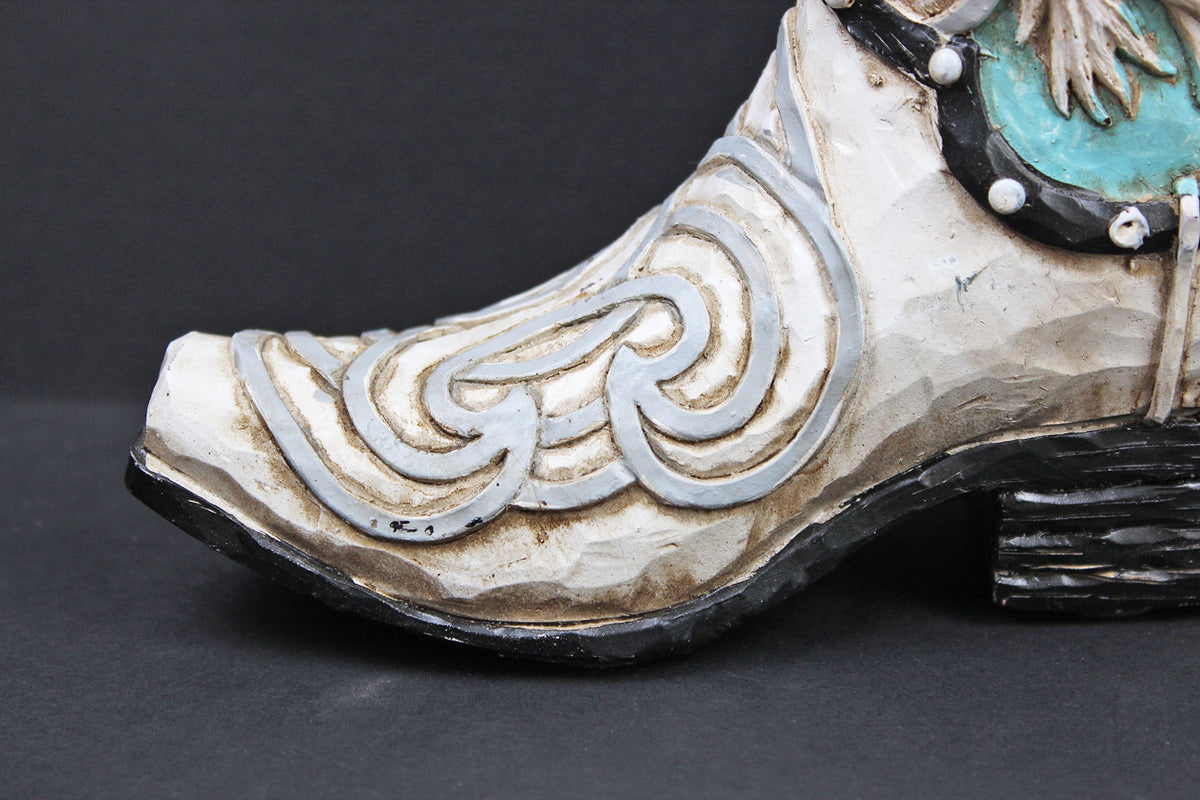 A close-up of a decorative cowboy boot featuring a distressed off-white leather base with silver-toned scrollwork, a turquoise inlay, and black detailing on a dark background.
