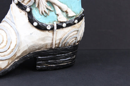 A close-up of a western-style boot with a distressed cream-colored leather upper, black heel and trim, turquoise blue painted detailing, and white decorative accents.