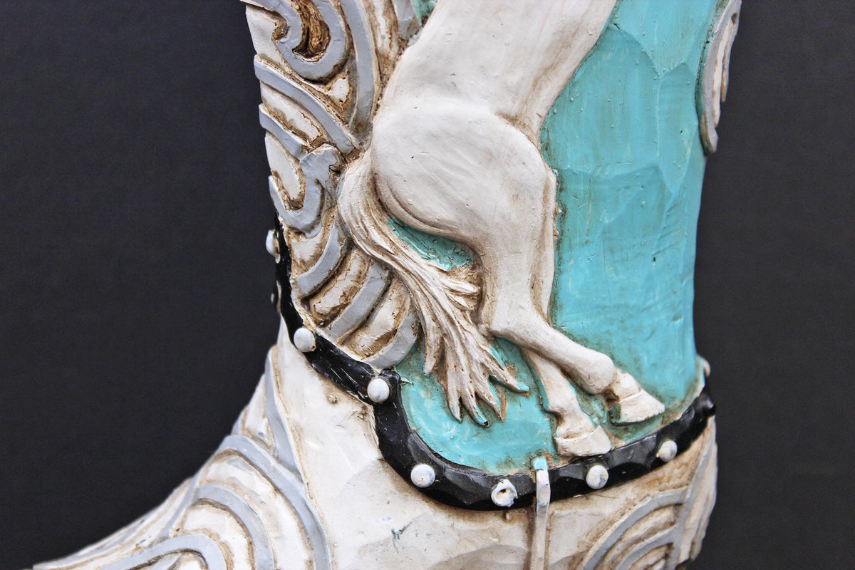 Close-up of a western-style boot featuring a sculpted white horse design over a turquoise blue inlay, with black and silver decorative trim and small rhinestone accents, set against a dark background.