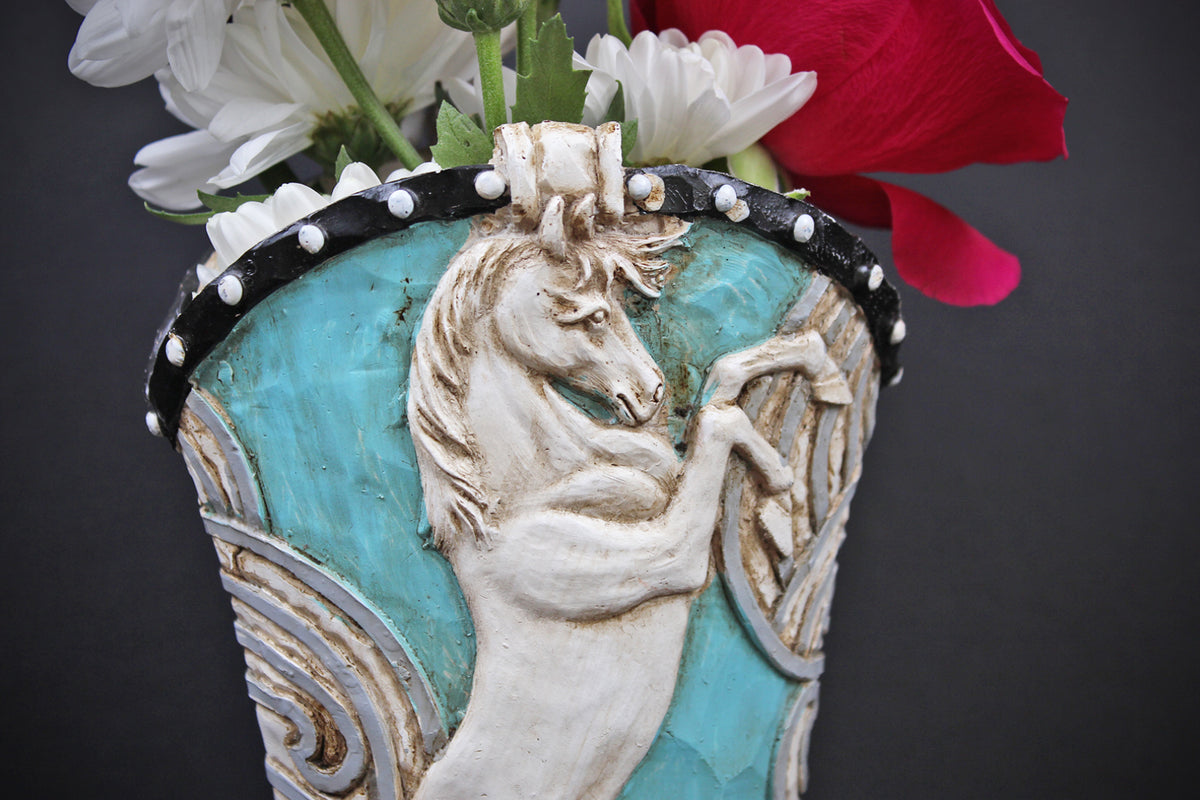 A decorative ceramic vase featuring a raised white horse motif on a turquoise blue background, with a black rim accented by white beads, holding white and red flowers against a dark gray backdrop.