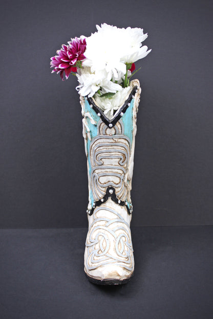 A decorative ceramic cowboy boot vase filled with white and purple flowers, set against a dark gray background.