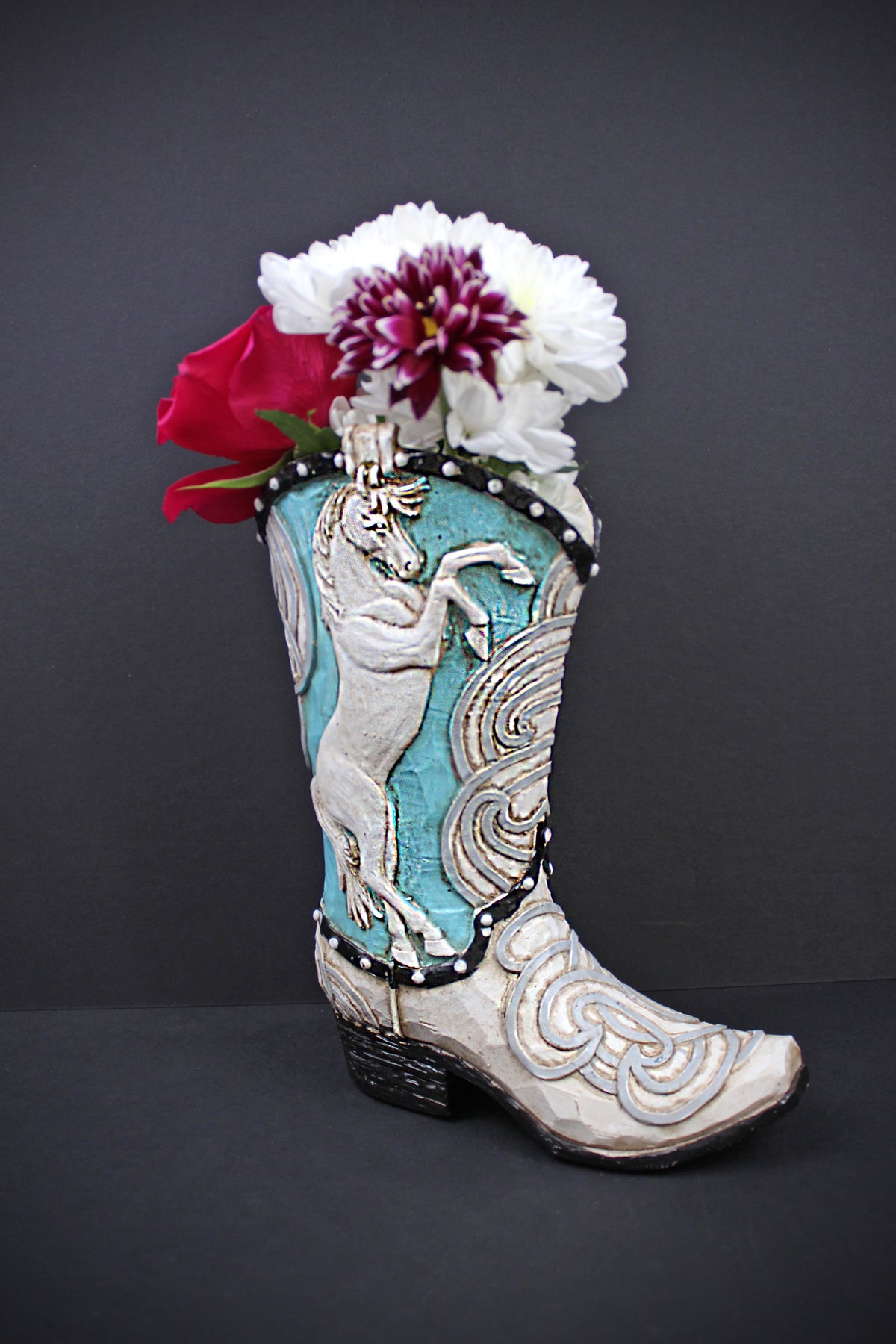 A decorative cowboy boot-shaped planter with turquoise and white western embroidery, filled with white and red flowers, set against a dark gray background.
