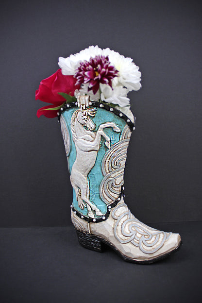 A decorative cowboy boot-shaped planter with turquoise and white western embroidery, filled with white and red flowers, set against a dark gray background.