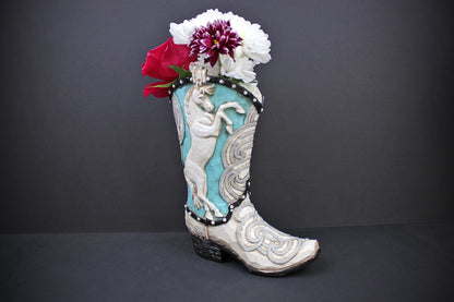 A decorative cowboy boot-shaped planter with turquoise and white western embroidery, holding white and red flowers, set against a dark gray background.
