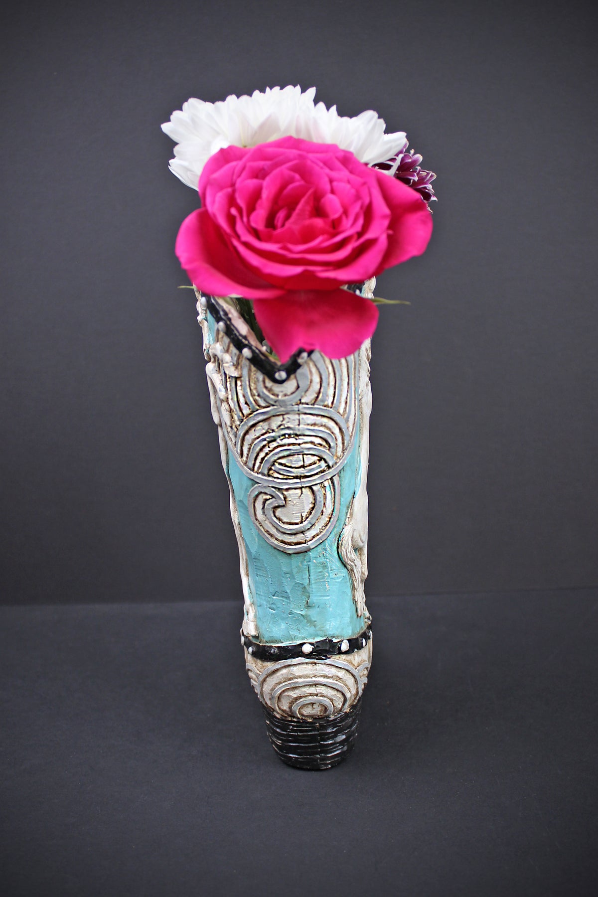 A ceramic vase shaped like a western boot, featuring turquoise blue and white textured patterns, filled with a bright pink rose and a white flower against a dark gray background.