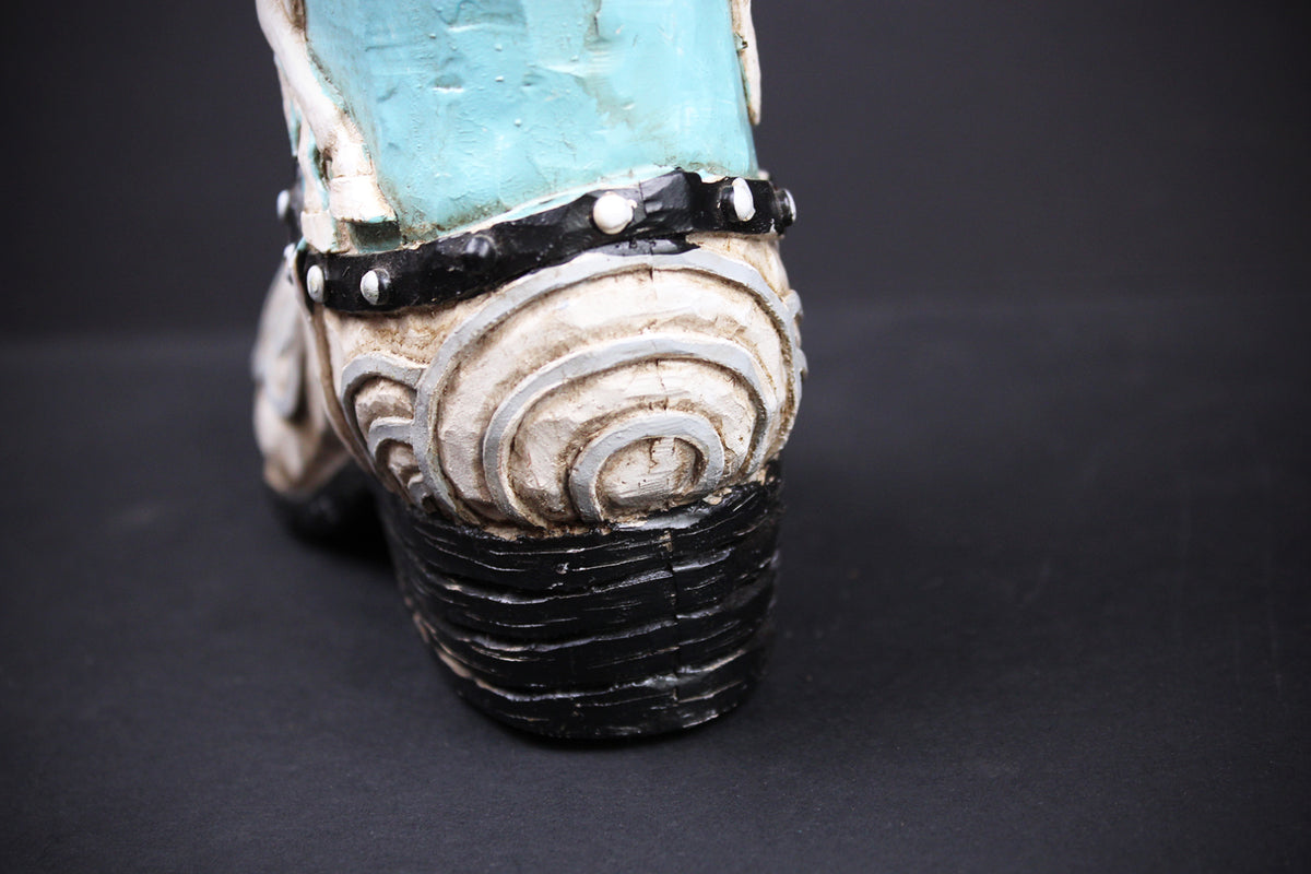 A close-up shot of a western-style boot heel, showing a distressed turquoise blue upper, a black studded strap, and a carved cream-colored spiral pattern on the heel counter, set against a dark background.
