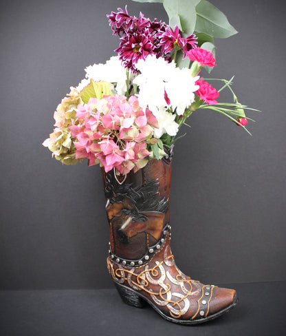A brown western-style cowboy boot with carved floral details and silver studs, filled with a bouquet of pink, white, and burgundy flowers, set against a dark gray background.