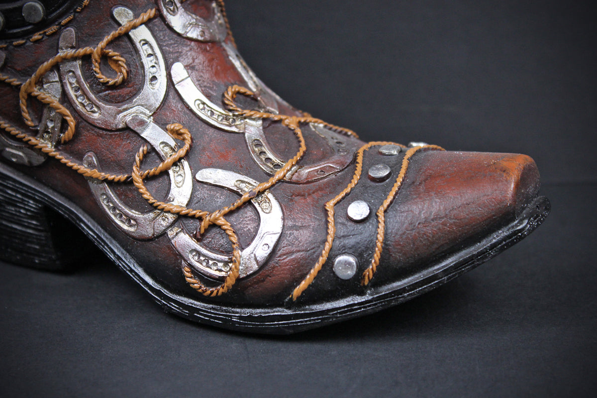 A close-up of a brown leather cowboy boot with silver horseshoe-shaped metal embellishments, tan rope laces, and silver studs on a dark background.