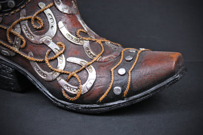 A close-up of a brown leather cowboy boot with silver horseshoe-shaped metal embellishments, tan rope laces, and silver studs on a dark background.