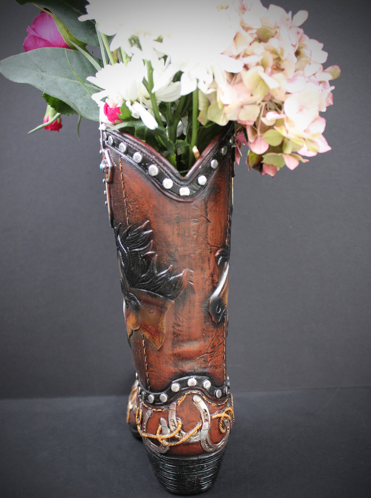 A brown leather cowboy boot vase filled with white and pink flowers, set against a dark gray background.