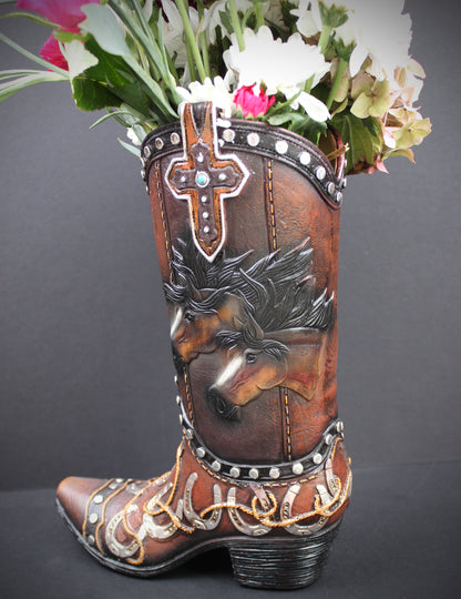 A brown leather cowboy boot vase filled with white and pink flowers, featuring hand-tooled western designs including a horse head, a cross, and silver and gold accents, set against a dark gray background.