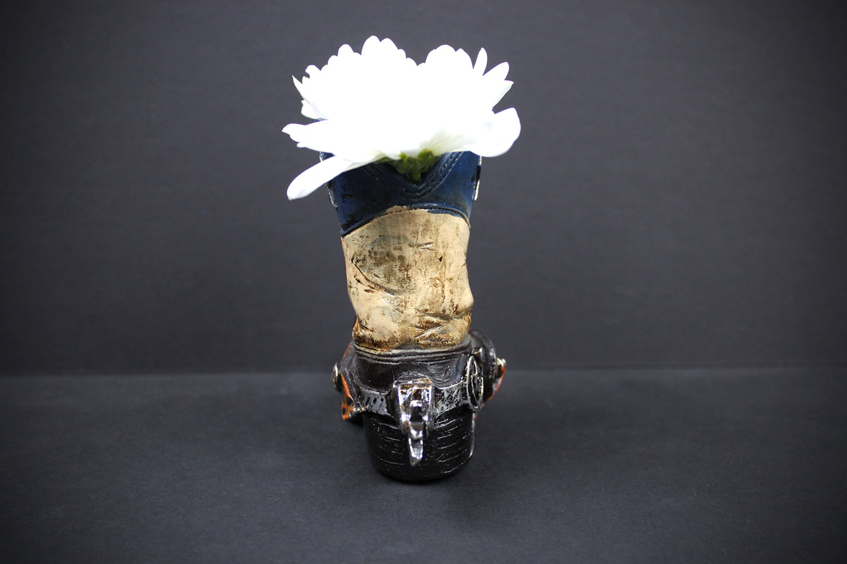 A gothic-style high-heeled shoe vase decorated with a blue denim upper, a weathered wood midsection, and a black faux-leather heel, holding a white flower against a dark gray background.