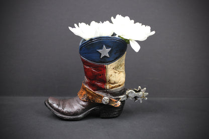 A decorative cowboy boot-shaped planter holding white flowers, set against a dark gray background. The boot features a distressed Texas flag design with blue, red, and gold panels, a silver star, and a brown leather-textured foot with a silver spur.
