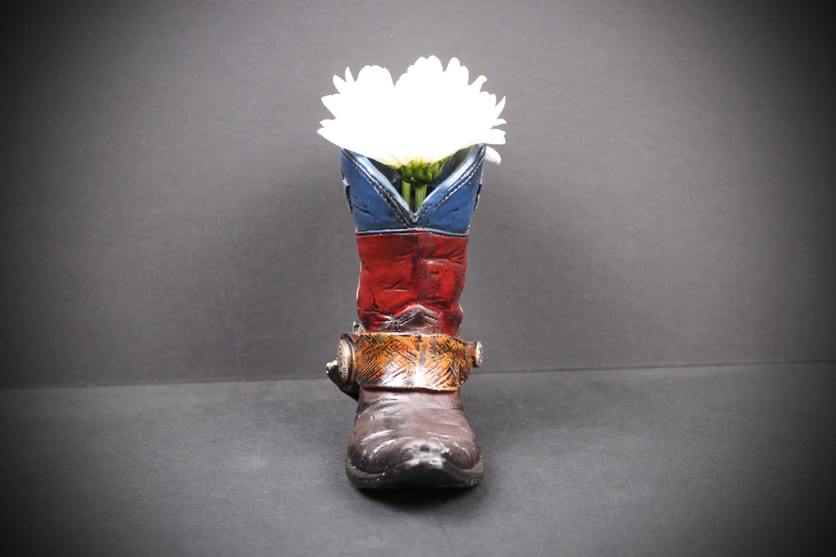 A decorative cowboy boot-shaped planter with a white flower inserted into the opening, set against a dark gray background.