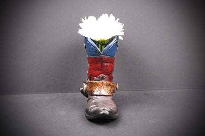 A decorative cowboy boot-shaped planter with a white flower inserted into the opening, set against a dark gray background.