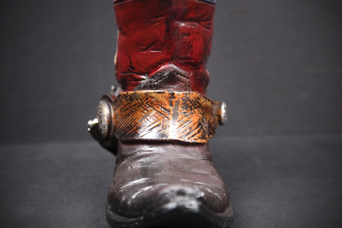 A close-up of a western-style boot with a dark brown leather foot, a wide metallic bronze-toned buckle strap, and a red leather shaft, set against a dark gray background.